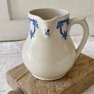 Puede incluir: Una jarra de cerámica blanca con diseños florales azules. La jarra tiene un cuerpo redondeado, un asa y un pico. Está sobre una tabla de cortar de madera. Es un artículo vintage o antiguo.