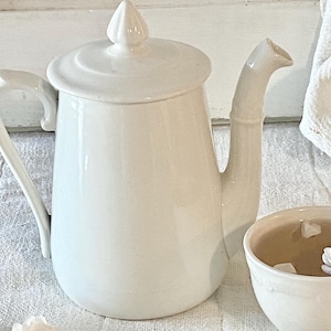 以下が含まれることがあります： クリーム色のセラミックティーポットと、それに合う蓋、そして紅茶の入ったカップ。 ティーポットは長い注ぎ口とハンドルが付いています。 カップには紅茶と白い花が入っています。 白いバラの花びらと花が散らばっています。