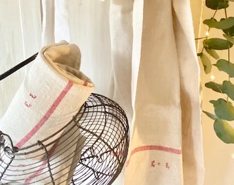 May include: A white linen tea towel with red stripes and embroidered details hangs on a white wooden rack. A wire basket with a rolled up linen tea towel is hanging below.