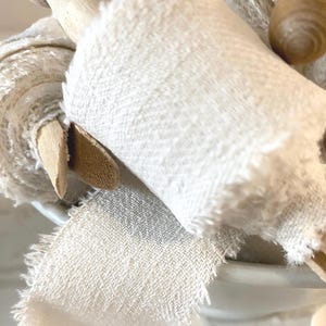 Könnte beinhalten: Nahaufnahme einer Rolle cremefarbenen Stoffes mit ausgefranstem Rand, möglicherweise Leinen oder Baumwolle. Eine hölzerne Teigrolle ist teilweise sichtbar, was auf ein Bastel- oder Wohnkulturthema hindeutet. Der Stoff hat ein strukturiertes Gewebe.