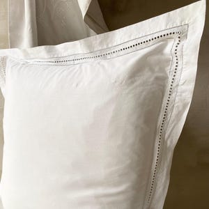May include: A white square pillow with a decorative border featuring small, evenly spaced cut-out details. The pillow is made of a soft, smooth fabric and is part of a bedding set.