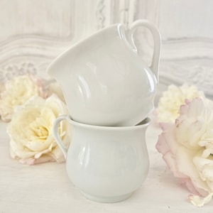 Op de afbeelding: Twee witte keramische theekopjes die op elkaar gestapeld zijn. De kopjes hebben een afgeronde vorm en een handvat aan één kant. De kopjes staan op een wit oppervlak met witte bloemen op de achtergrond.