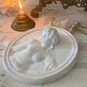 May include: A white plaster relief sculpture of a reclining figure in an oval frame. The sculpture is on a distressed white surface, with a lit candle in a brass holder and a decorative brass candelabra in the background. The theme is classical art.
