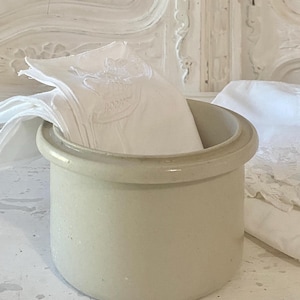 May include: A beige ceramic crock with a white rim, sitting on a white surface. White fabric napkins are stacked inside the crock.