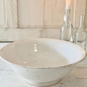 Peut inclure: Un grand bol en c&eacute;ramique blanc cass&eacute; avec un large bord et un socle. Le bol est vide et pos&eacute; sur une surface blanche vieillie. En arri&egrave;re-plan, il y a deux bouteilles en verre transparent et une bougie allum&eacute;e dans une bouteille.