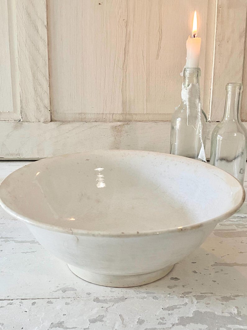 Peut inclure: Un grand bol en c&eacute;ramique blanc cass&eacute; avec un large bord et un socle. Le bol est vide et pos&eacute; sur une surface blanche vieillie. En arri&egrave;re-plan, il y a deux bouteilles en verre transparent et une bougie allum&eacute;e dans une bouteille.