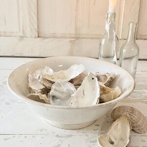Peut inclure: Un bol en c&eacute;ramique blanc rempli de coquilles d'hu&icirc;tres. Deux bouteilles en verre transparent et une bougie allum&eacute;e dans une bouteille sont en arri&egrave;re-plan. Les coquilles sont blanc cass&eacute; et beiges.