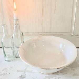 Peut inclure: Un bol en c&eacute;ramique blanc au bord l&eacute;g&egrave;rement irr&eacute;gulier est pos&eacute; sur une surface en bois blanc vieilli. Deux bouteilles en verre transparent et une bougie allum&eacute;e dans une bouteille sont &agrave; gauche, cr&eacute;ant une esth&eacute;tique rustique et vintage.