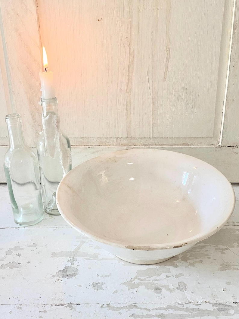 Peut inclure: Un bol en c&eacute;ramique blanc au bord l&eacute;g&egrave;rement irr&eacute;gulier est pos&eacute; sur une surface en bois blanc vieilli. Deux bouteilles en verre transparent et une bougie allum&eacute;e dans une bouteille sont &agrave; gauche, cr&eacute;ant une esth&eacute;tique rustique et vintage.
