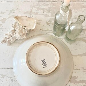 Peut inclure: Une assiette en c&eacute;ramique blanche avec un bord marron, le dessous affichant un embl&egrave;me noir et blanc et le texte "DIGOIN FRANCE". L'image pr&eacute;sente &eacute;galement deux bouteilles en verre transparent, une bougie allum&eacute;e, une coquille d'hu&icirc;tre et un morceau de corail sur une surface en bois blanc.