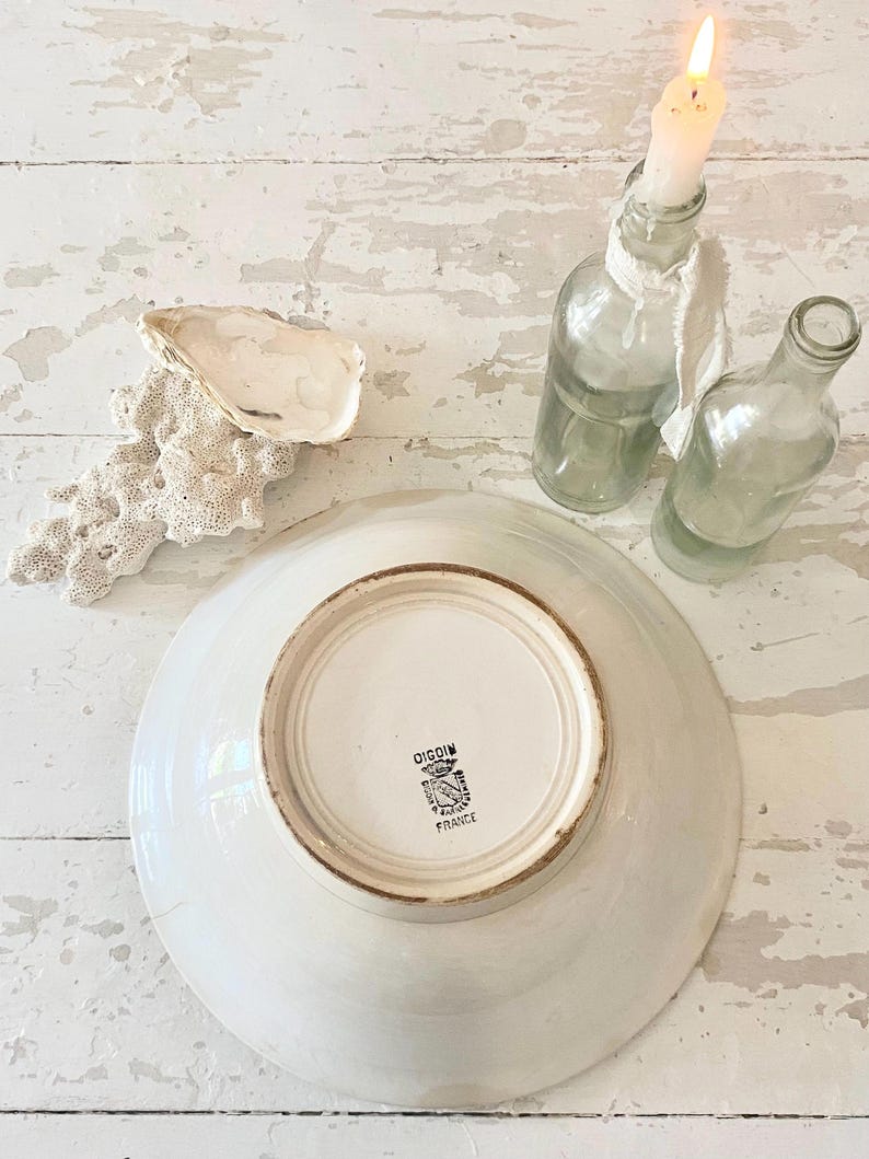 Peut inclure: Une assiette en c&eacute;ramique blanche avec un bord marron, le dessous affichant un embl&egrave;me noir et blanc et le texte "DIGOIN FRANCE". L'image pr&eacute;sente &eacute;galement deux bouteilles en verre transparent, une bougie allum&eacute;e, une coquille d'hu&icirc;tre et un morceau de corail sur une surface en bois blanc.