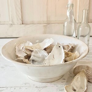 Peut inclure: Un bol en c&eacute;ramique blanc rempli de coquilles d'hu&icirc;tres. Les coquilles sont de diff&eacute;rentes nuances de beige et de blanc. Deux bouteilles en verre transparent et une bougie allum&eacute;e dans une bouteille sont en arri&egrave;re-plan.