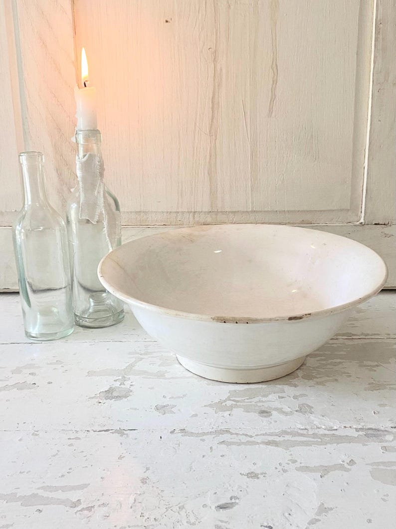 Peut inclure: Un grand bol en c&eacute;ramique blanche avec un large bord repose sur une surface en bois blanc vieilli. Deux bouteilles en verre transparent et une bougie allum&eacute;e dans une bouteille en verre sont derri&egrave;re le bol. La bougie a une flamme vive.