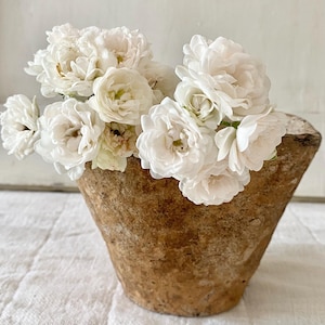 Op de afbeelding: Een bruine, kegelvormige vaas gevuld met een boeket witte rozen. De rozen hebben meerdere lagen delicate bloemblaadjes. De vaas staat op een wit, getextureerd oppervlak, wat een contrast creëert tussen de ruwe textuur van de vaas en de zachte bloemen.