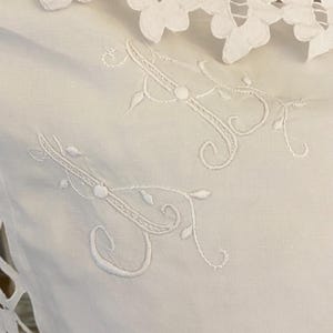 May include: A white textile item featuring intricate white floral cutwork lace along the edges and delicate white embroidered monogram letters in a cursive script style, showcasing detailed needlework on a smooth, solid white fabric surface.
