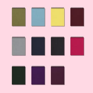 Puede incluir: Una colección de doce cuadernos con espiral en varios colores, incluyendo verde oliva, azul claro, amarillo y burdeos. Los cuadernos están dispuestos sobre un fondo rosa claro, mostrando sus diversos tonos y texturas.