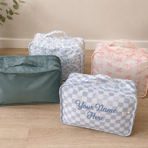 May include: Four rectangular zippered travel pouches with top handles. One is solid sage green, one has a blue floral pattern, one features a pink bow print, and one has a blue and white checkerboard pattern with the text Your Name Here.