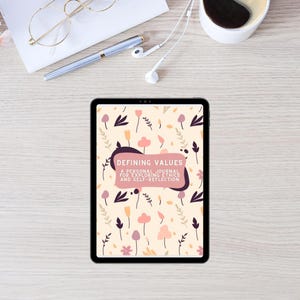 May include: A digital tablet displaying a journal cover with the text "Defining Values: A Personal Journal for Exploring Ethics and Self-Reflection." The cover features a floral pattern in pink, purple, and beige tones. Other items include glasses, a pen, and headphones.
