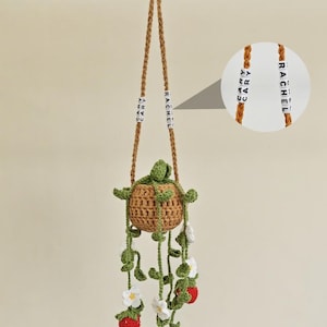 May include: A handcrafted crochet hanging planter, featuring a brown woven basket with green leaves and red strawberries. The planter is suspended by two brown cords with the names "CARY" and "RACHEL" spelled out in white beads. White flowers are also included.