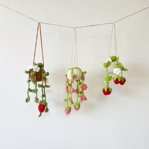 Może przedstawiać: Trzy wiszące doniczki wykonane na szydełku z kwiatami i truskawkami. Doniczki są brązowe, białe i białe z różowymi kwiatami. Truskawki są czerwone.