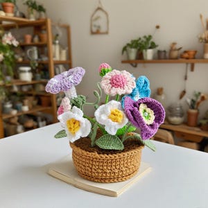 Puede incluir: Un arreglo floral de ganchillo en una maceta marrón. El arreglo incluye flores blancas, rosas, moradas y azules con hojas verdes. La maceta está sobre un libro. El fondo incluye estanterías y plantas.