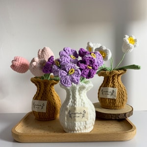May include: Three crocheted vases containing a bouquet of crocheted flowers. The vases are white, brown and beige. The flowers are purple, pink and white. The vases have a label that says "Handmade with Love".