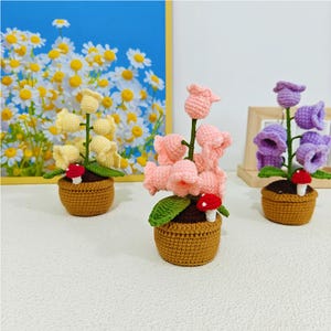 May include: Three crocheted flower arrangements in pots. The flowers are pink, purple, and yellow. Each pot has a small red mushroom.