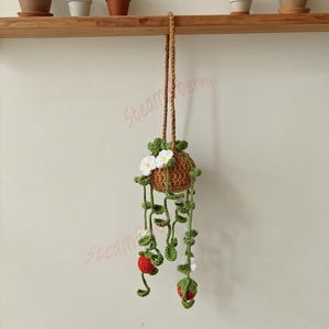 May include: A handmade crochet hanging planter featuring a brown basket, green vines, white flowers, and red berries. The planter is suspended by a brown rope, designed for small plants. Decorative and whimsical.