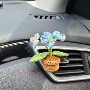May include: A crocheted flower in a small brown pot, designed to be placed in a car's air vent. The flower has blue, pink, and green petals and green leaves. The pot is a light brown color.