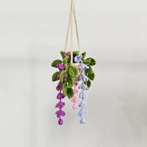Może przedstawiać: Ręcznie robiona, szydełkowa donica wisząca z pnączami i kwiatami. Donica jest beżowa, z zielonymi liśćmi i fioletowymi, różowymi i niebieskimi kwiatami. Sznurek do zawieszania jest w naturalnym beżowym kolorze.
