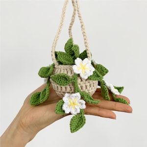 May include: A crocheted hanging planter with white flowers and green leaves. The planter is made of beige yarn and has a simple, rustic look.