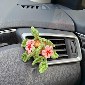 May include: Hand-crocheted car air vent decoration. The design features a turtle with green leaves, a tan shell, and pink flowers with red centers. The accessory is attached to a car's air vent.