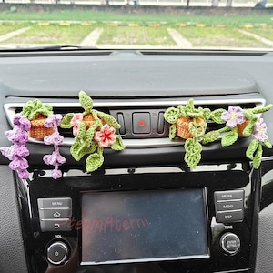 May include: Crocheted car vent decorations shaped like potted plants. The plants have green leaves and colourful flowers in shades of purple, pink, and orange. The crocheted pots are brown.