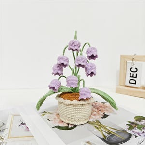 Puede incluir: Una planta de lirio de los valles de crochet en una maceta blanca con una base marrón. La planta tiene hojas verde claro y flores moradas.