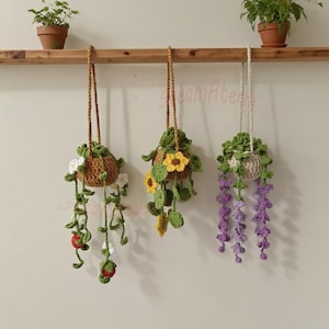 May include: Three handmade crochet hanging planters with floral designs. The planters are brown, green, white, yellow, and purple. They hang from a wooden beam. The crocheted plants feature detailed craftsmanship.