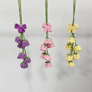 Könnte beinhalten: Drei gehäkelte Blumengirlanden in Lila, Rosa und Gelb. Jede Girlande hat eine Schleife oben und eine Gruppe glockenförmiger Blumen, die an einem grünen Stiel hängen. Der Hintergrund ist schlicht weiß.