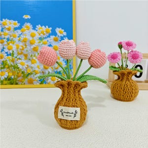 May include: Two crocheted flower arrangements in brown pots. The first arrangement features pink crocheted tulips in a brown pot with a "Handmade with Love" tag. The second arrangement features pink crocheted flowers in a brown pot.
