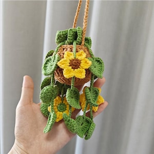 May include: A crocheted sunflower hanging planter with green leaves and a brown pot. The planter is made of yarn and has a yellow sunflower with a brown centre.