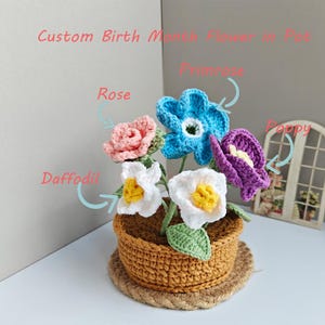 Könnte beinhalten: Handgefertigtes Häkelblumenarrangement in einem braunen Topf. Das Arrangement umfasst eine rosa Rose, blaue Primel, weiße Narzisse und lila Mohnblume. Der Topf steht auf einem gewebten Untersetzer. Text: "Custom Birth Month Flower in Pot."