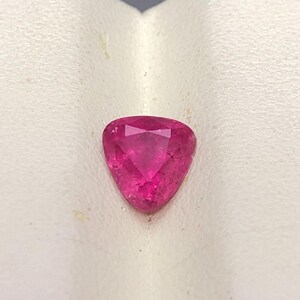May include: A vibrant, deep pink gemstone cut into a triangular shape with multiple facets. The stone displays a brilliant, translucent quality with visible internal inclusions and sharp, defined edges forming a classic trilliant cut silhouette.