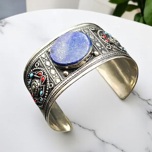 Può includere: Bracciale a polsino in argento con una gemma ovale blu al centro. Il bracciale presenta un'intricata lavorazione del metallo con accenti rossi e turchesi. Il design è ornato e dettagliato, in stile vintage o etnico.