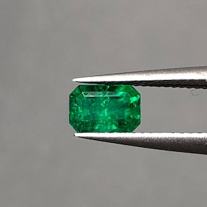 May include: A vibrant, rectangular-cut emerald gemstone held by tweezers. The gem is a deep, rich green color, with visible facets and internal clarity. The emerald is displayed against a neutral gray background, highlighting its brilliance.