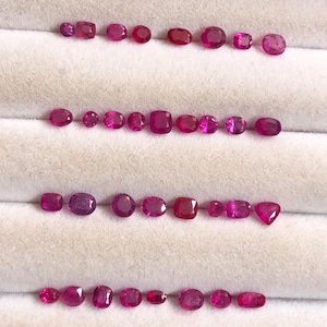May include: A collection of loose, faceted rubies in various shapes and sizes, including oval, cushion, and round cuts. These vibrant red gemstones are arranged in four horizontal rows, showcasing deep pinkish-red hues and reflective surfaces.