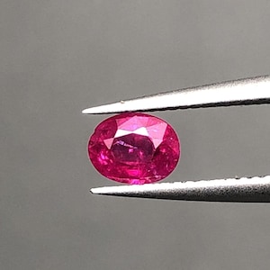May include: A vivid, oval-cut ruby gemstone held by tweezers against a grey backdrop. The ruby exhibits a deep, saturated red hue with facets that reflect light, highlighting its brilliance. The tweezers are silver.
