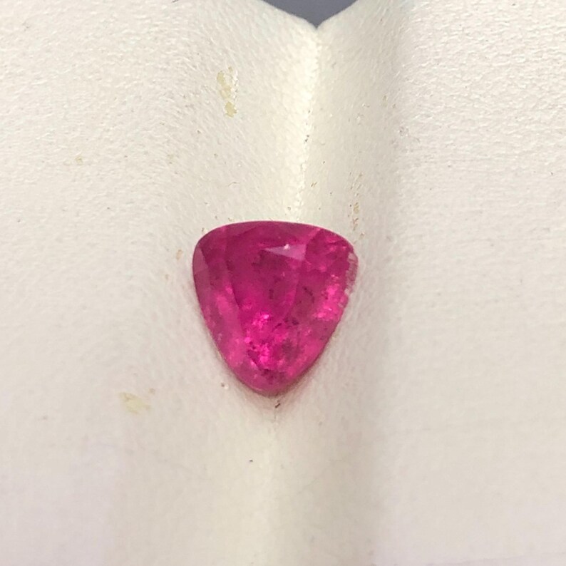 May include: A loose, faceted gemstone with a vibrant hot pink color and a triangular shape. The stone features multiple polished surfaces that reflect light, creating a brilliant appearance across its top face and pointed bottom edges.