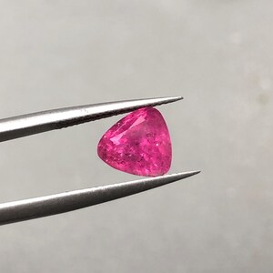 May include: A vibrant, hot pink gemstone with a triangular, faceted cut held by silver-toned metal tweezers. The stone displays internal clarity and light-reflecting surfaces, showcasing a deep magenta hue throughout its translucent, geometric structure.