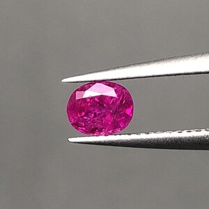 May include: A faceted, oval-cut ruby gemstone held by tweezers. The ruby is a vibrant shade of pinkish-red, with visible internal inclusions. The tweezers are silver-colored, and the background is a solid gray.