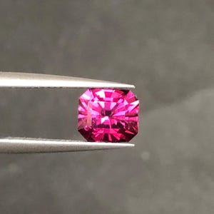 Puede incluir: Una radiante gema facetada de corte cuadrado en un vibrante tono magenta. La gema es sostenida por unas pinzas plateadas sobre un fondo gris oscuro, mostrando su intrincada estructura interna y brillo. Ideal para joyería.