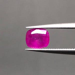 Könnte beinhalten: Ein facettierter Rubin im Kissenschliff, gehalten von einer Pinzette. Der Rubin hat eine lebendige, tiefrosa-rote Farbe mit einer polierten Oberfläche, die das Licht reflektiert. Die Form des Edelsteins ist ein abgerundetes Quadrat.