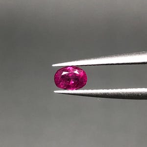 May include: A vibrant, oval-cut gemstone with a deep magenta-pink hue and multifaceted surface, held securely between the tips of a pair of silver-toned metal tweezers. The stone displays internal clarity and light reflection across its polished facets.